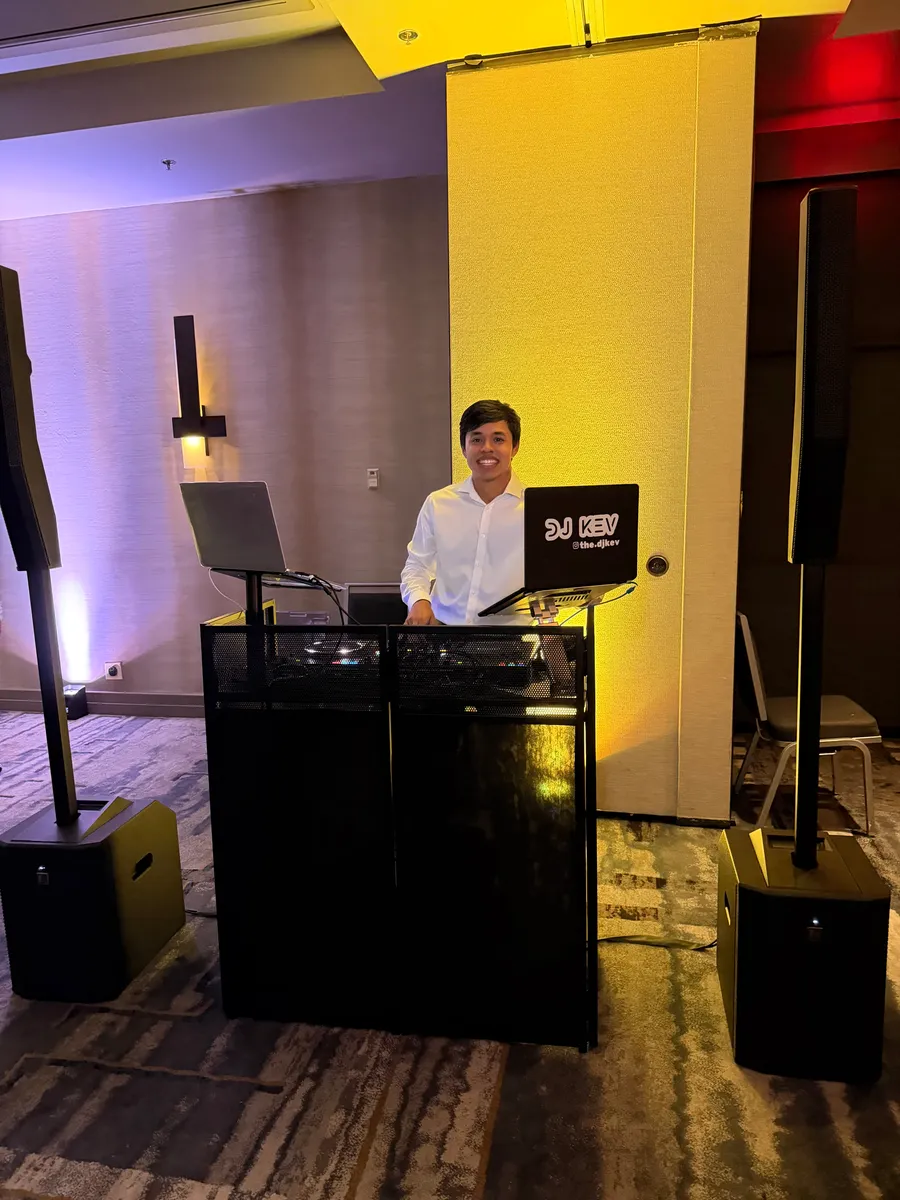 DJ Kev smiling behind his DJ booth and speakers set up in a hotel ballroom
